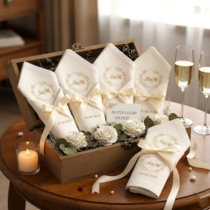May include: A wooden box filled with folded white napkins, each adorned with gold embroidery of "A&M" and the date "20.06.2024". The napkins are tied with cream-colored ribbons. The box also contains white roses and greenery, with two champagne flutes in the background.