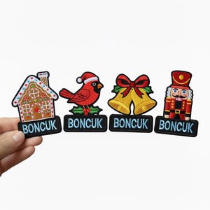 Könnte beinhalten: Gestickte Weihnachtspatches mit einem Lebkuchenhaus, einem roten Kardinal mit Weihnachtsmütze, goldenen Glocken mit roter Schleife und einem Spielzeugsoldaten. Jeder Patch hat das Wort "BONCUK" auf schwarzem Hintergrund.