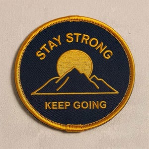 May include: A circular embroidered patch with a dark navy blue background and a gold border. The design features a gold sun rising behind a mountain range, with the text STAY STRONG arched above and the text KEEP GOING printed below the mountains.