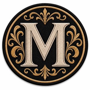 Op de afbeelding: Een ronde geborduurde patch met een grote, crèmekleurige letter M in het midden op een zwarte achtergrond. De letter is omringd door ingewikkelde, symmetrische goudkleurige krullen en bloemmotieven, alles omsloten door een dunne gouden rand.