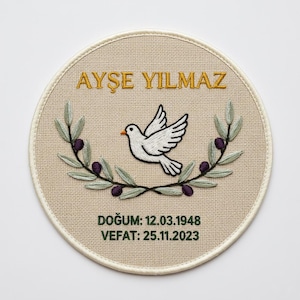 Può includere: Pezzo commemorativo rotondo ricamato con il nome "AYŞE YILMAZ" in lettere dorate. Una colomba bianca è centrata sopra un ramo d'ulivo verde con olive viola. Il testo "DOĞUM: 12.03.1948 VEFAT: 25.11.2023" è sotto.