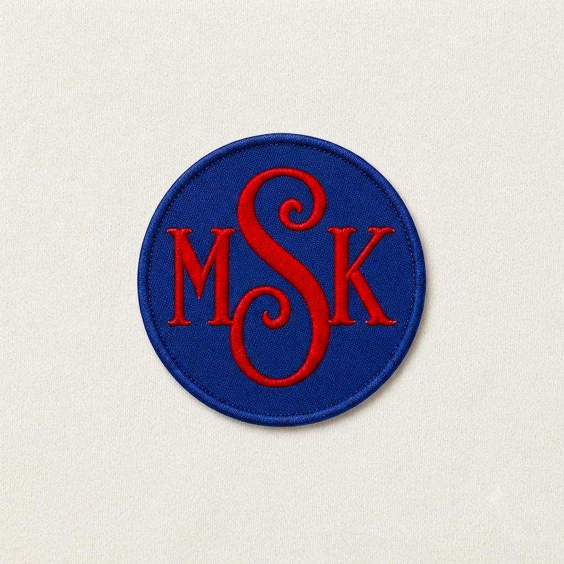 May include: A circular embroidered patch featuring a royal blue background with a red monogram in the centre. The monogram consists of the letters M, S, and K, with the central letter S being larger and more ornate than the letters M and K.