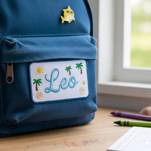 Op de afbeelding: Een blauwe rugzak met een patch met de naam "Leo" in cursiefschrift, palmbomen, een zon en schelpen. Er is een gele haaienpin bevestigd. De rugzak heeft een voorvak met een rits.