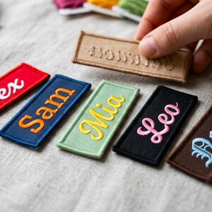 Puede incluir: Parches bordados con nombres en rojo, azul, verde, negro y marrón. Cada parche muestra un nombre diferente en un color contrastante, como blanco, naranja, amarillo y rosa. Una mano sostiene un parche beige.