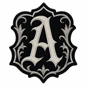Op de afbeelding: Een zwarte geborduurde patch in de vorm van een schild met een grote, witte, gotische letter A in het midden. De patch is versierd met ingewikkelde witte krullen en florale filigraanpatronen langs de randen van de zwarte achtergrond.