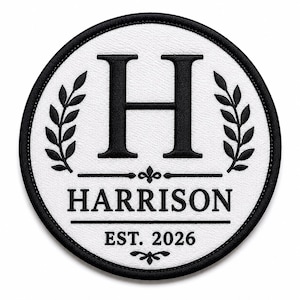 Op de afbeelding: Een ronde geborduurde patch met een witte achtergrond en een zwarte rand. Het bevat een grote zwarte letter H, twee zwarte lauriertakken, de tekst HARRISON en de tekst EST. 2026, allemaal in zwart borduurwerk op een witte stoffen basis.