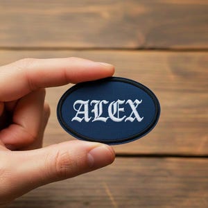 Op de afbeelding: Een ovaal marineblauw embleem met de naam "ALEX" geborduurd in witte, gotische letters. Het embleem heeft een zwarte rand en wordt in een hand gehouden tegen een houten achtergrond.