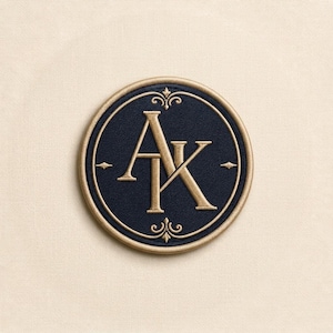 Op de afbeelding: Een ronde geborduurde patch met een donkerblauw midden en de goudkleurige letters AK in elkaar verstrengeld in het midden. De patch is omlijst door een gouden rand en bevat kleine decoratieve gouden versieringen aan de boven- en onderkant.