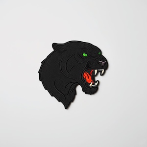 Könnte beinhalten: Ein schwarzer Panther-Aufnäher mit grünen Augen und offenem Maul, das ein rotes Innere und weiße Zähne zeigt. Der Aufnäher ist mit einem strukturierten Aussehen bestickt, und der Panther blickt nach links.