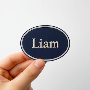 Può includere: Toppa ovale blu navy con il nome "Liam" ricamato in filo beige chiaro. La toppa ha un bordo bianco ed è presentata su sfondo bianco.