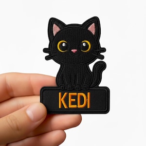 Könnte beinhalten: Schwarzer, gestickter Aufnäher mit einer Cartoon-Katze mit gelben Augen und rosa Ohren. Die Katze sitzt über einem rechteckigen schwarzen Banner mit dem Namen "KEDI" in orangefarbenen Buchstaben. Ein dekorativer Aufnäher für Katzenliebhaber.