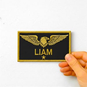 Könnte beinhalten: Schwarzer rechteckiger Aufnäher mit goldgesticktem Adlerdesign und dem Namen "LIAM" in goldfarbenen Buchstaben. Unter dem Namen befindet sich ein goldener Stern. Der Aufnäher hat einen goldenen Rand.