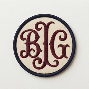 Puede incluir: Un parche bordado circular con un fondo de color crema y un borde cosido en azul marino oscuro. En el centro, las letras B, I y G se muestran en una fuente cursiva grande, ornamentada y de color burdeos oscuro con remolinos decorativos.