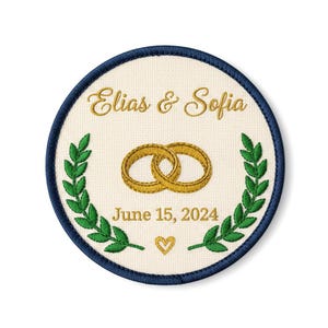 Puede incluir: Parche bordado circular con borde azul marino y centro crema. Presenta texto dorado "Elias & Sofia" sobre dos anillos dorados entrelazados. Debajo, "June 15, 2024" y un corazón dorado.