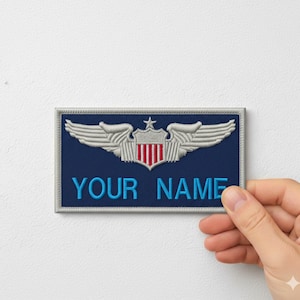 May include: A navy blue embroidered patch with the text "YOUR NAME" in bright blue. The patch features a silver emblem with wings, a star, and a shield with red and white stripes.