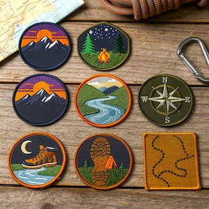 Puede incluir: Parches bordados con temas de exteriores, incluyendo montañas, una fogata, un río, una brújula, una bota de senderismo, una huella y un mapa. Los parches presentan varios colores como azul, naranja, verde y marrón, y están dispuestos sobre una superficie de madera.