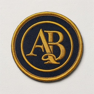 May include: A circular embroidered patch featuring a dark navy blue background with a gold border. In the center, the letters A and B are intertwined in a gold serif font, enclosed within a thin gold circular line that matches the outer edge.