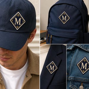 May include: A navy blue baseball cap, a navy blue backpack, a navy blue blazer, and a blue denim jacket, each featuring a diamond-shaped patch with a cream-coloured border and a central letter M in a serif font, accented with small decorative flourishes.