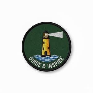 Op de afbeelding: Geborduurde patch met een gele vuurtoren, zwart dak en witte lichtstraal op een donkergroene achtergrond. De patch heeft de witte woorden "GUIDE & INSPIRE" binnen een zwarte cirkelvormige rand.