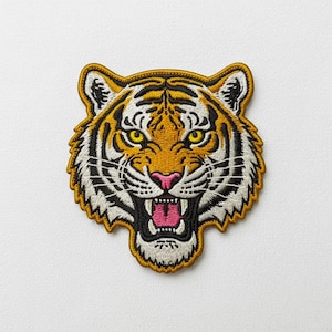 Peut inclure: Un écusson brodé représentant une tête de tigre avec des yeux jaunes, un nez rose et une gueule ouverte montrant des dents et une langue rose. Le tigre présente des motifs de fourrure orange, noirs et blancs avec une bordure dorée épaisse.