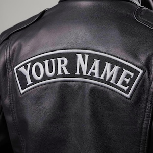 Può includere: Giacca in pelle nera con una toppa ricamata bianca con la scritta "YOUR NAME" in un design curvo. La toppa è bordata con cuciture bianche ed è cucita sul retro della giacca. La giacca ha un classico stile biker.
