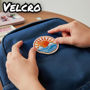 Pu&ograve; includere: Una toppa ricamata circolare raffigurante un sole arancione con raggi radianti sopra onde oceaniche blu e bianche. La toppa &egrave; posizionata su una superficie in tessuto denim blu scuro. La parola VELCRO &egrave; stampata in grandi lettere bianche.
