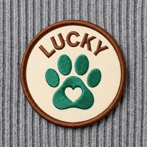 Pode incluir: Um emblema bordado circular com fundo creme e borda marrom. O emblema exibe a palavra LUCKY em letras marrons acima de um ícone de pegada verde que contém um pequeno recorte em formato de coração no centro.