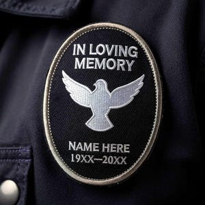 May include: A black patch with white embroidered text and a dove design. The text reads "IN LOVING MEMORY", "NAME HERE", and dates. The patch is oval-shaped with a silver border and is sewn onto a dark blue fabric.