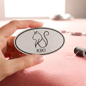 Könnte beinhalten: Graues, ovales Namensschild mit schwarzem Rand und dem Namen "KIKI" unter einer Katzen-Silhouette. Das Schild wird von einer Hand gehalten, im Hintergrund eine Garnrolle und eine Schere. Das Schild ist wahrscheinlich für ein Haustier.