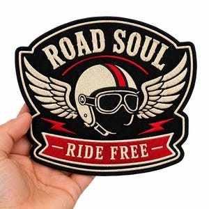 Op de afbeelding: Een zwarte geborduurde patch met een crèmekleurige motorhelm met een rode streep en een bril, geflankeerd door vleugels. De tekst ROAD SOUL staat in een boog erboven en RIDE FREE staat op een rode banner eronder, met twee rode bliksemschichten.
