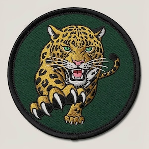 Könnte beinhalten: Gestickter Aufnäher mit einem detaillierten Jaguar-Design. Der Jaguar hat ein gelb-schwarz geflecktes Fell, grüne Augen und eine rosa Nase. Der Aufnäher ist kreisförmig, mit einem dunkelgrünen Hintergrund und einem schwarzen Rand.