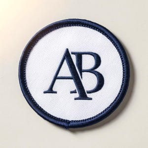 Op de afbeelding: Geborduurde ronde patch met de marineblauwe letters "AB" op een witte achtergrond. De patch heeft een marineblauwe gestikte rand. Deze patch kan worden gebruikt op kleding of accessoires. Het ontwerp is eenvoudig en elegant.