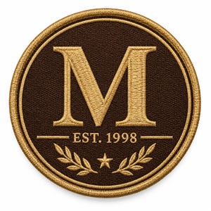 Puede incluir: Un parche bordado circular con fondo marrón oscuro y borde dorado. Una letra M dorada grande está centrada en la parte superior, con el texto EST. 1998 debajo, flanqueado por dos líneas horizontales, una pequeña estrella y ramas de laurel.