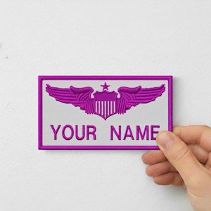 May include: A rectangular patch with a white background and a magenta border. The patch features a magenta emblem of wings and a shield with a star. The text "YOUR NAME" is embroidered in magenta.