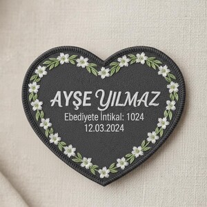 Puede incluir: Un parche en forma de corazón con fondo gris oscuro, bordado con flores blancas y hojas verdes en el borde. El nombre "AYSE YILMAZ" está bordado en blanco, junto con texto adicional y una fecha.