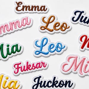 May include: A collection of embroidered name patches featuring various names including Emma, Leo, Juma, Mia, Fuksar, and Juckon. Each patch is rendered in a cursive script font with a white border, showcasing diverse thread colors like pink, green, blue, yellow, brown, and dark navy.
