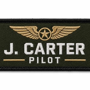 Può includere: Una toppa rettangolare ricamata color verde oliva con bordo nero. Presenta un emblema dorato ricamato con una stella alata nella parte superiore. Sotto l'emblema, il testo J. CARTER è ricamato in bianco, seguito dalla parola PILOT.
