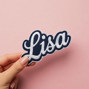 Pode incluir: Um patch bordado azul marinho com o nome "Lisa" em letras brancas. O patch tem uma borda branca e é mantido contra um fundo rosa.