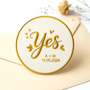 Puede incluir: Parche circular bordado con la palabra "Yes" en letras doradas, rodeado de un borde dorado. El parche incluye las iniciales "A + M" y la fecha "10.05.2024". El fondo es de color crema.