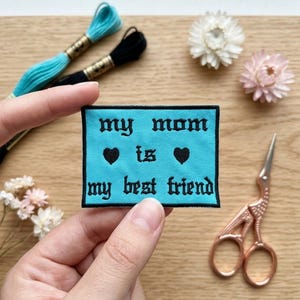 Może przedstawiać: Turkusowa naszywka z napisem "my mom is my best friend" czarną gotycką czcionką, z dwoma czarnymi symbolami serca. Naszywka trzymana jest w dłoni, z nicią do haftowania i małymi nożyczkami w tle.