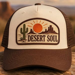 May include: A cream and brown trucker hat featuring an embroidered patch on the front. The patch displays a green cactus, an orange sun, brown mountains, and the text "DESERT SOUL" in dark brown lettering against a textured tan background.