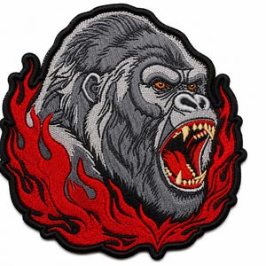 Könnte beinhalten: Ein gestickter Aufnäher mit einem grauen Gorillakopf mit offenem Maul und sichtbaren Zähnen, umgeben von stilisierten roten Flammen. Das Design ist mit schwarzem Garn umrandet und erzeugt ein kontrastreiches grafisches Element mit detaillierten Stichen.