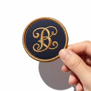 Op de afbeelding: Geborduurde patch met een marineblauwe achtergrond en een goudkleurige letter "B" in een decoratief schrift. De patch heeft een gouden rand en is rond van vorm. De patch wordt door een hand tegen een witte achtergrond gehouden.