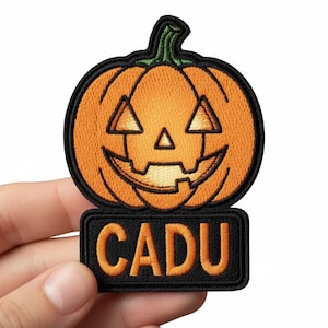 Op de afbeelding: Geborduurde patch met een lachende pompoen en het woord "CADU" eronder. De pompoen is oranje met een groene stengel en zwarte omtrek. De tekst is oranje op een zwarte achtergrond.