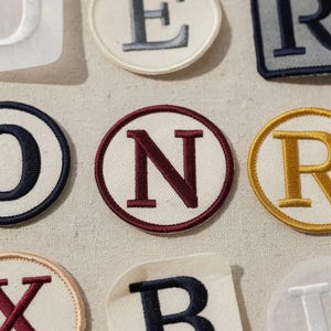 May include: Close-up of embroidered letter patches in various colors. The patches feature letters like "N" in burgundy, "R" in gold, and "B" in navy blue. The patches are on a neutral fabric background.