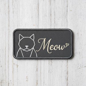 May include: A rectangular dark grey embroidered patch featuring a white geometric line art illustration of a cat head on the left side and the word Meow in white cursive script followed by a small heart outline on the right side.