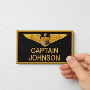 Könnte beinhalten: Ein schwarzer rechteckiger Aufnäher mit goldfarbenen Stickereien. Der Aufnäher zeigt ein goldenes Emblem aus Flügeln, einem Stern und einem Schild. Der Text "CAPTAIN JOHNSON" ist goldfarben gestickt. Der Aufnäher wird von einer Hand gehalten.