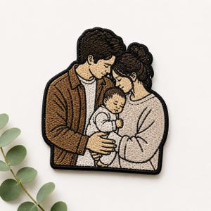 May include: An embroidered patch featuring a family of three. A person in a brown button-down shirt stands next to a person in a light beige jumper, both holding a baby dressed in white. The design uses black outlines on a tan and brown colour palette.