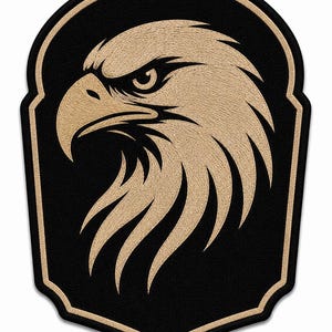 May include: A shield-shaped embroidered patch featuring a stylized eagle head in tan thread against a solid black background. The eagle is depicted in profile facing left with sharp, detailed feathers and a prominent beak, bordered by a tan edge.