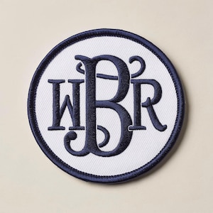 Könnte beinhalten: Runder, gestickter Aufnäher mit weißer Mitte und marineblauem Rand. Der Aufnäher zeigt das marineblaue Monogramm "WBR" in einer dekorativen Schriftart, geeignet zur Personalisierung von Kleidung oder Accessoires.
